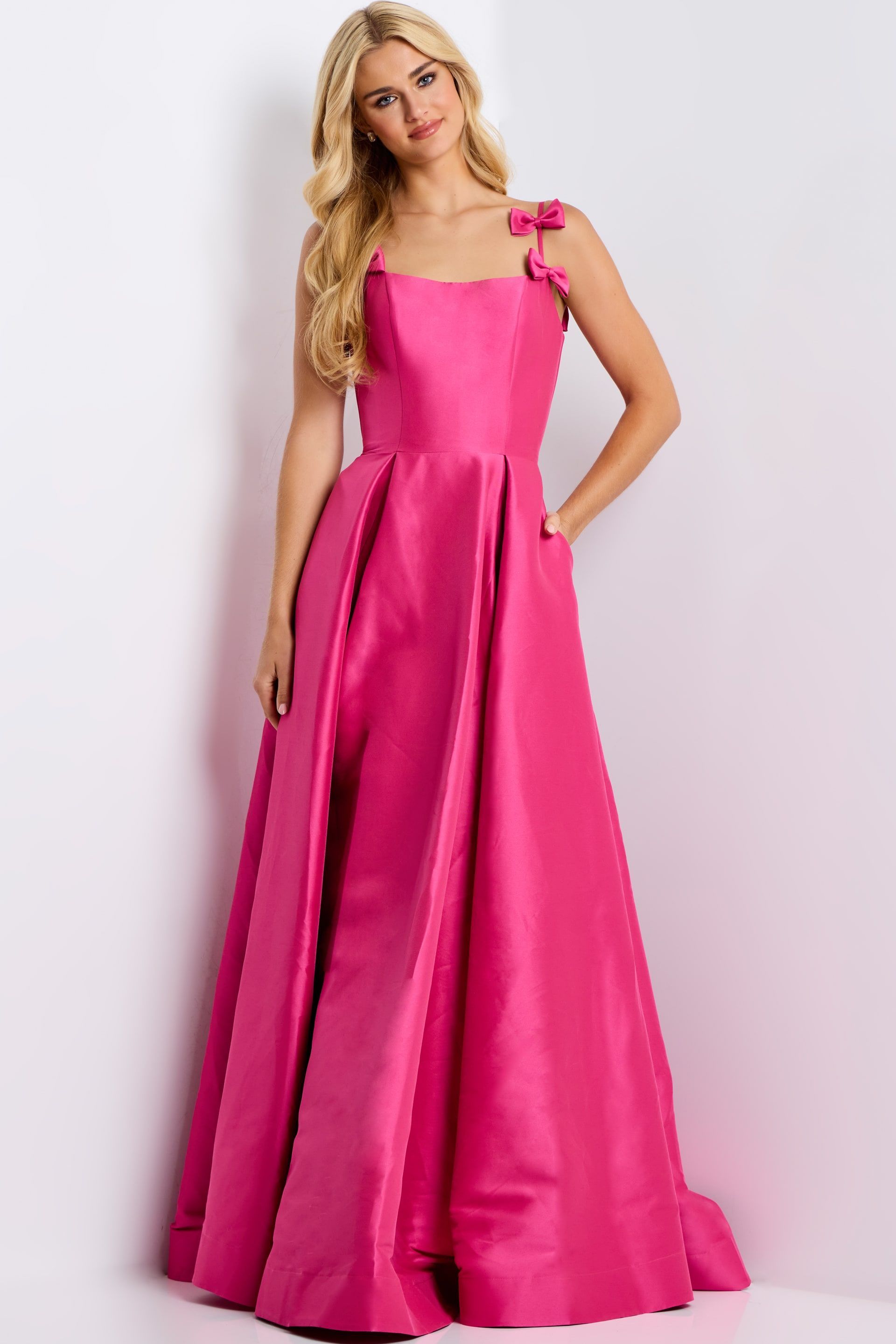 Woman in a bright pink gown, standing indoors with one hand in the pocket. The dress has bow details on straps.