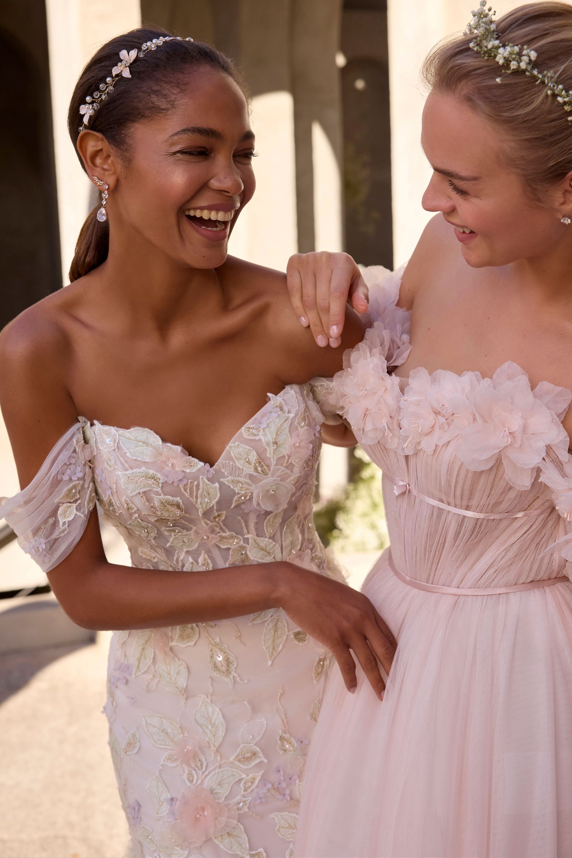 Two women in pastel dresses laughing, outdoors. One has a silver headband, the other a floral crown.