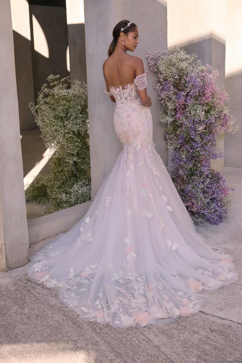 Woman in off-shoulder, mermaid-style wedding dress with floral detailing; stands near flowers in a bright setting.