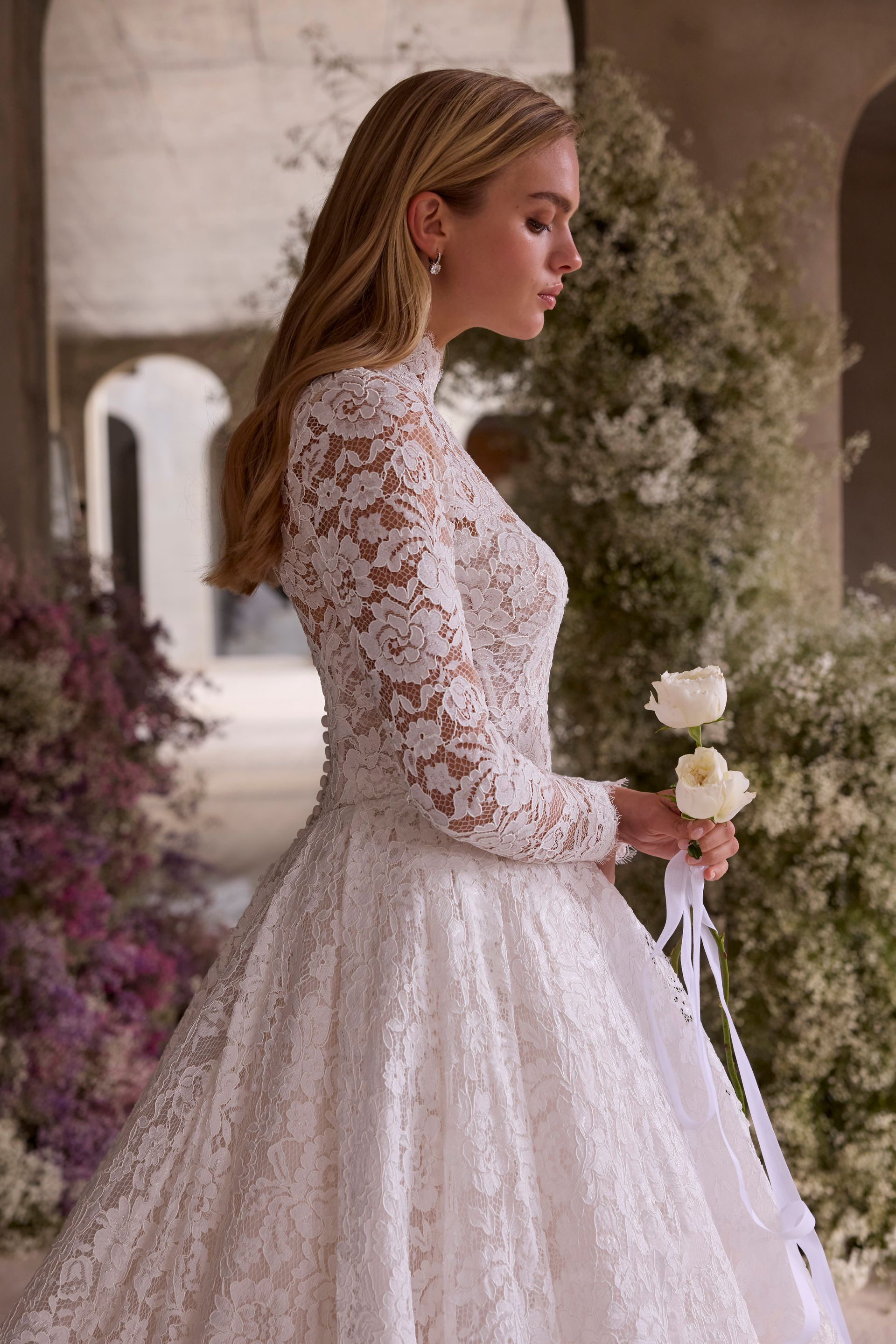 Blonde woman in a lace wedding dress holding white roses, standing outside near floral arrangements.