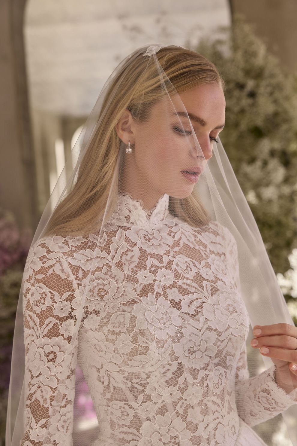Bride in white lace wedding dress, veil covers face, gazing down.