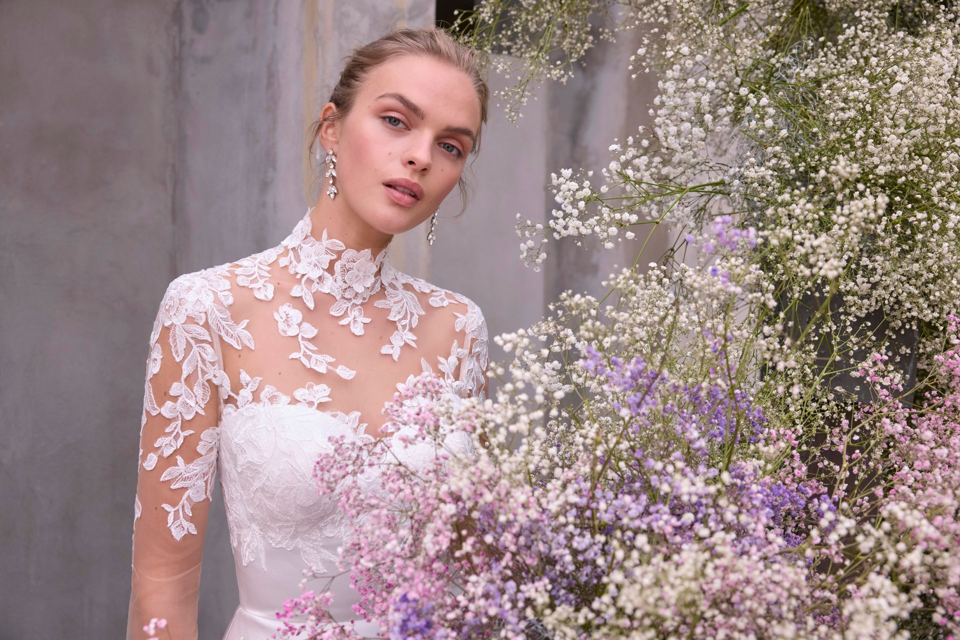 Woman in white lace wedding dress, surrounded by baby's breath flowers, against a gray wall.