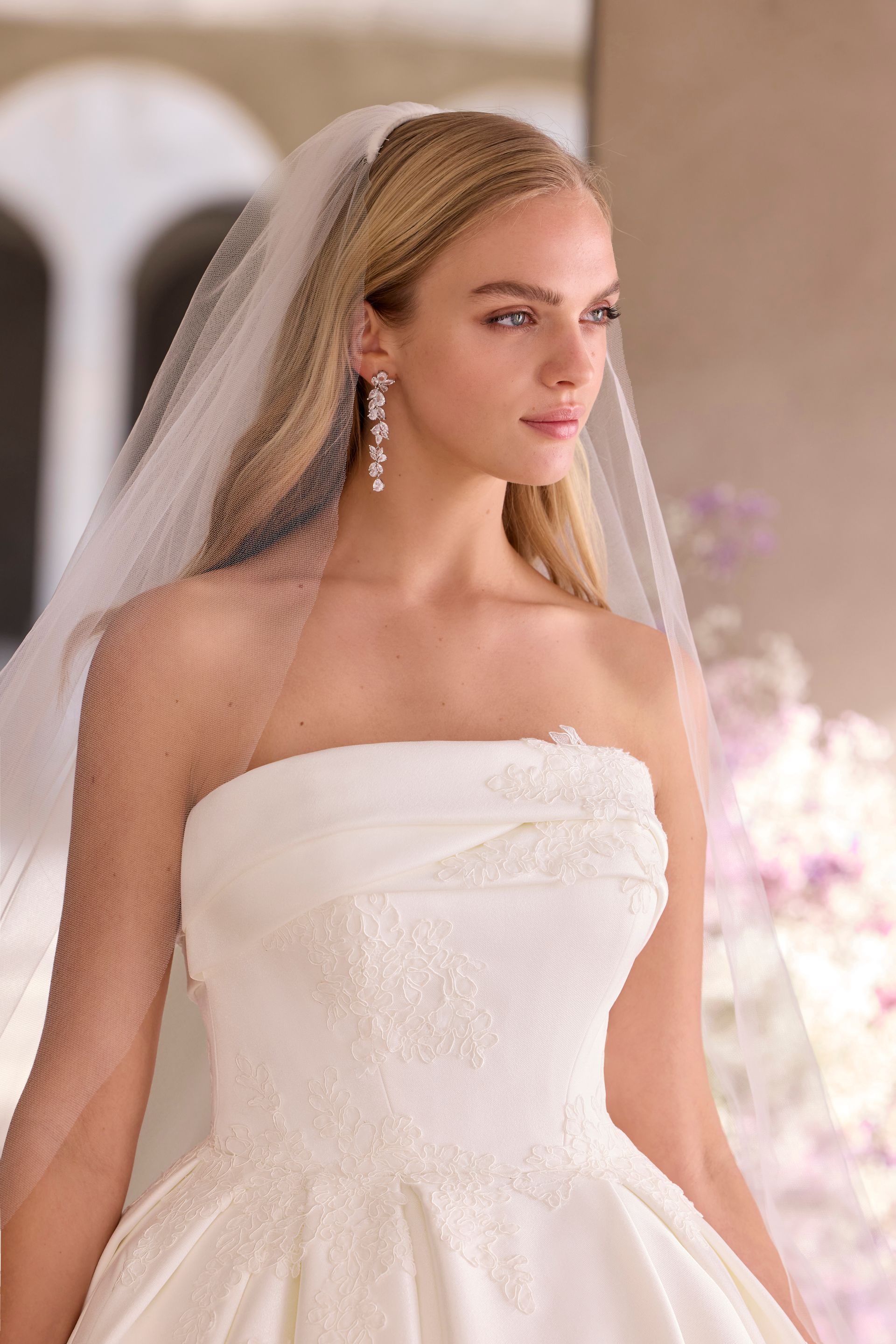 Bride in a white wedding dress and veil, holding white flowers, looking off to the side in soft light.