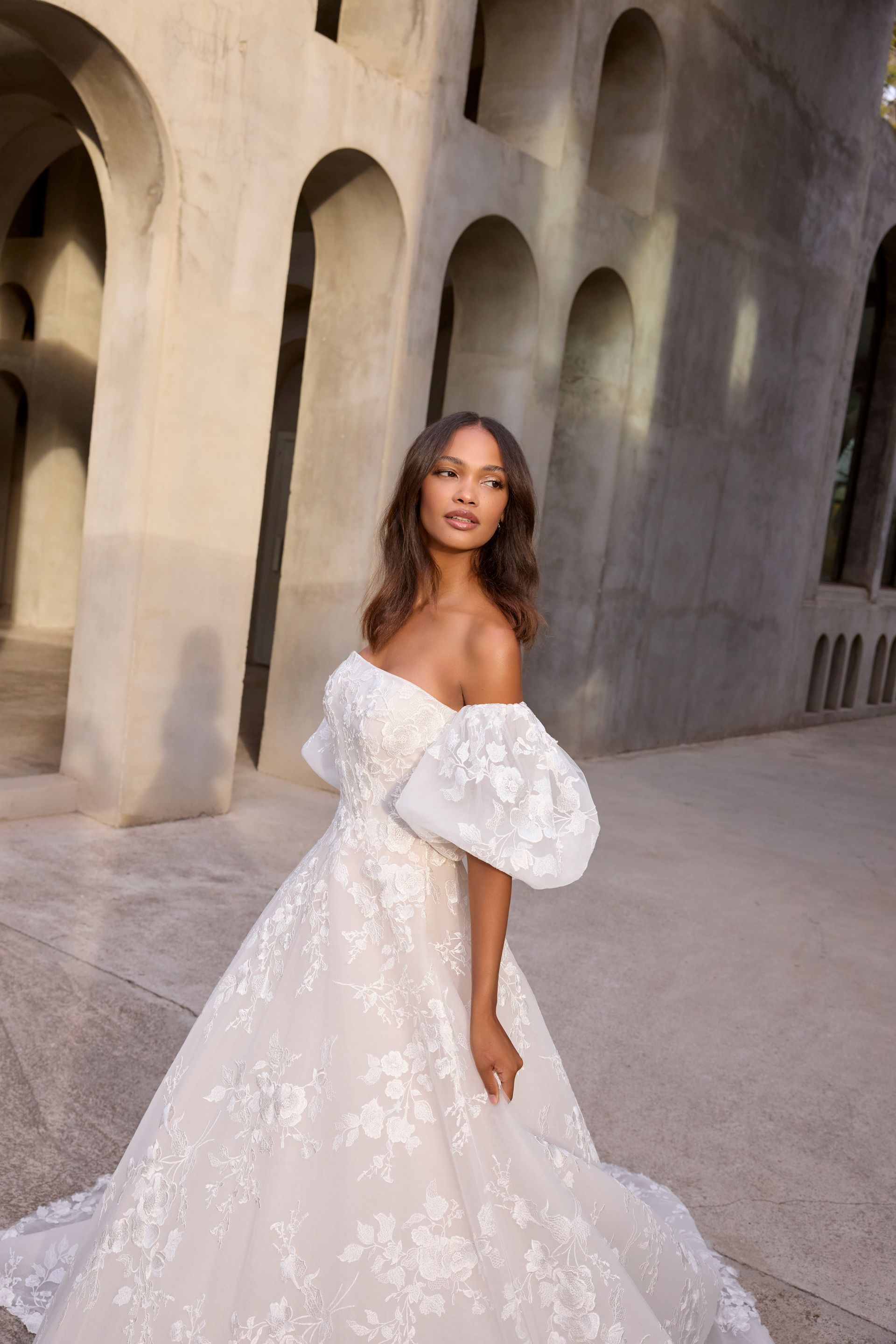 Woman in white off-the-shoulder wedding dress with puffy sleeves, standing outside by arched architecture.