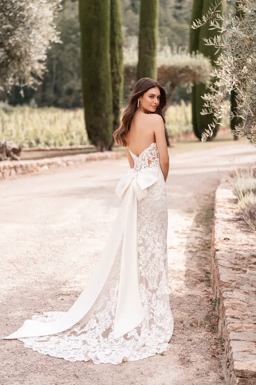 Woman in a white lace wedding dress with a large bow and train, standing on a path in a garden.