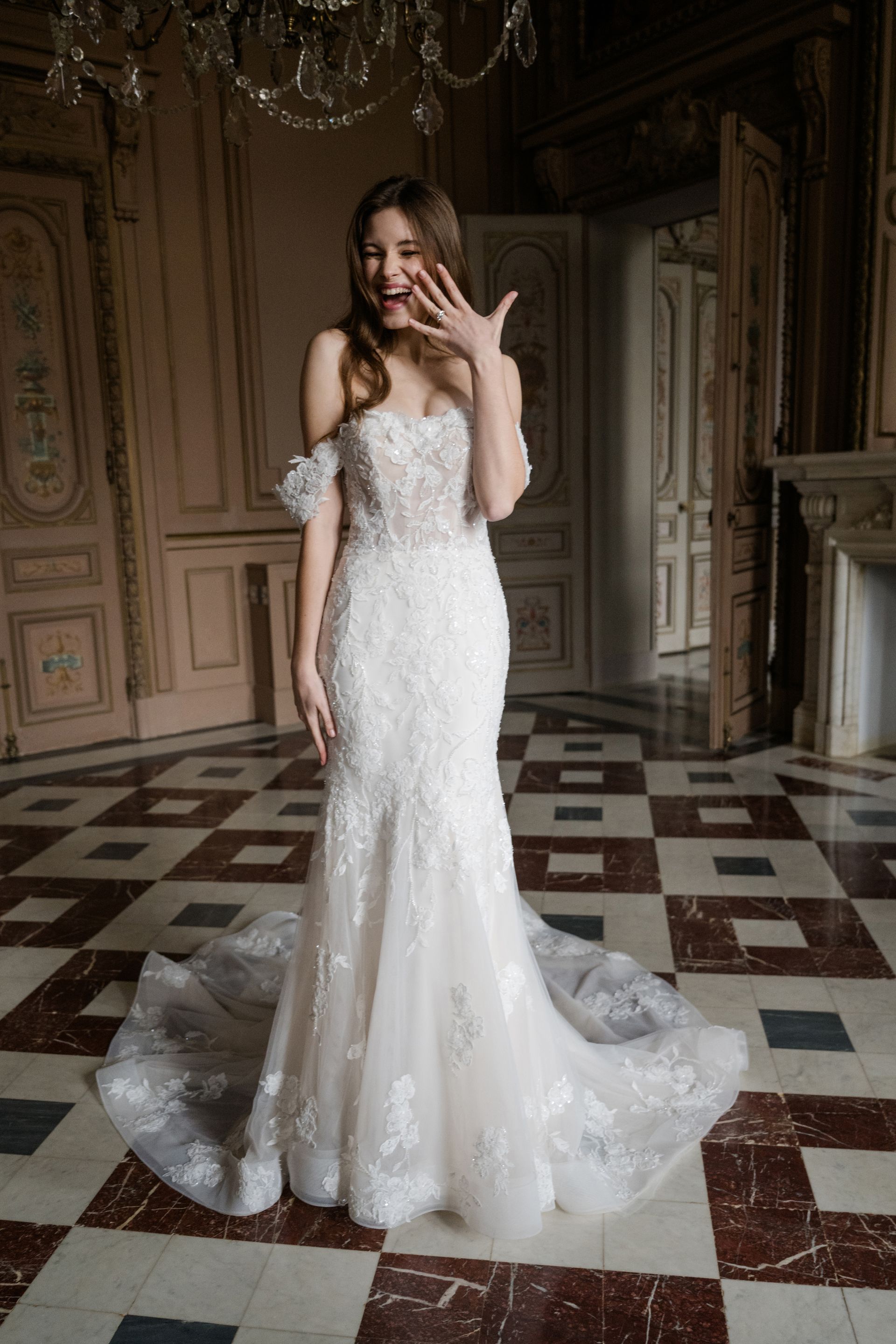 Bride in a white lace gown smiles, covering her mouth. She stands in a large room with patterned flooring.
