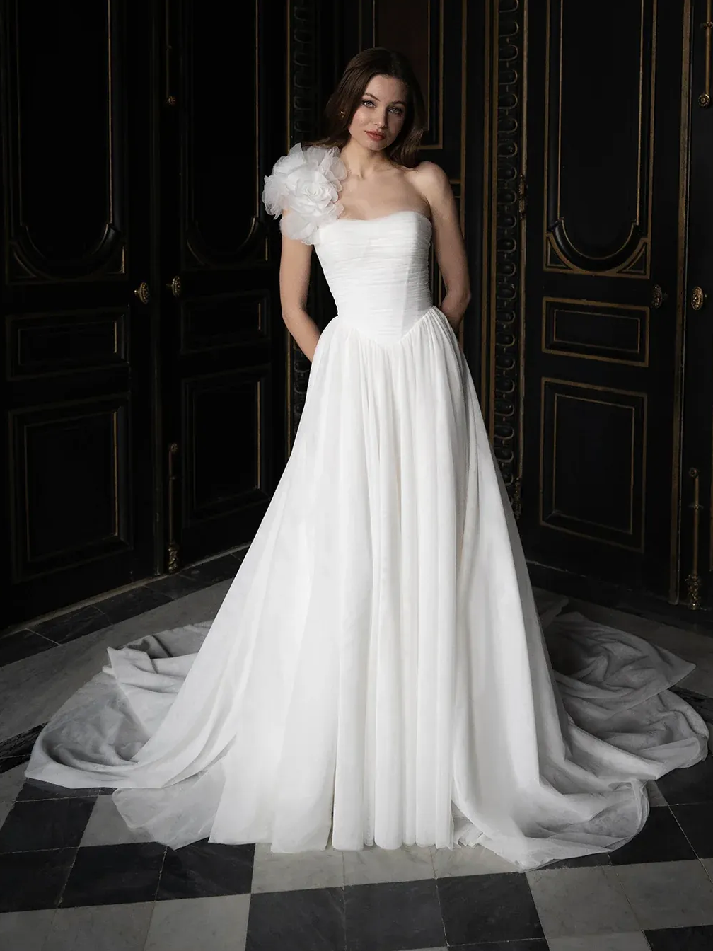 Woman in white, one-shoulder wedding dress, with floral accent, standing in front of ornate, dark doors.