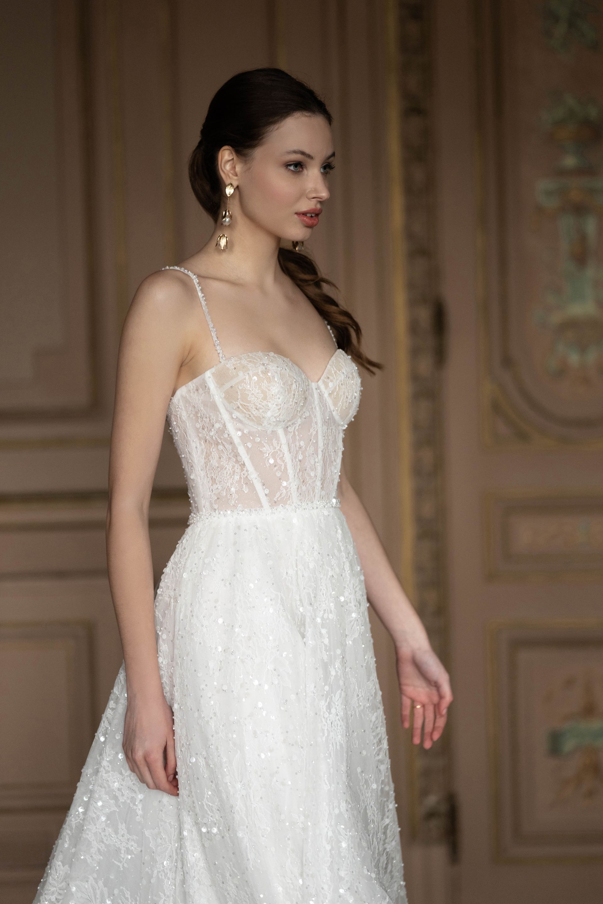 Woman in a beaded white wedding dress, side profile, standing indoors with wood panel doors.