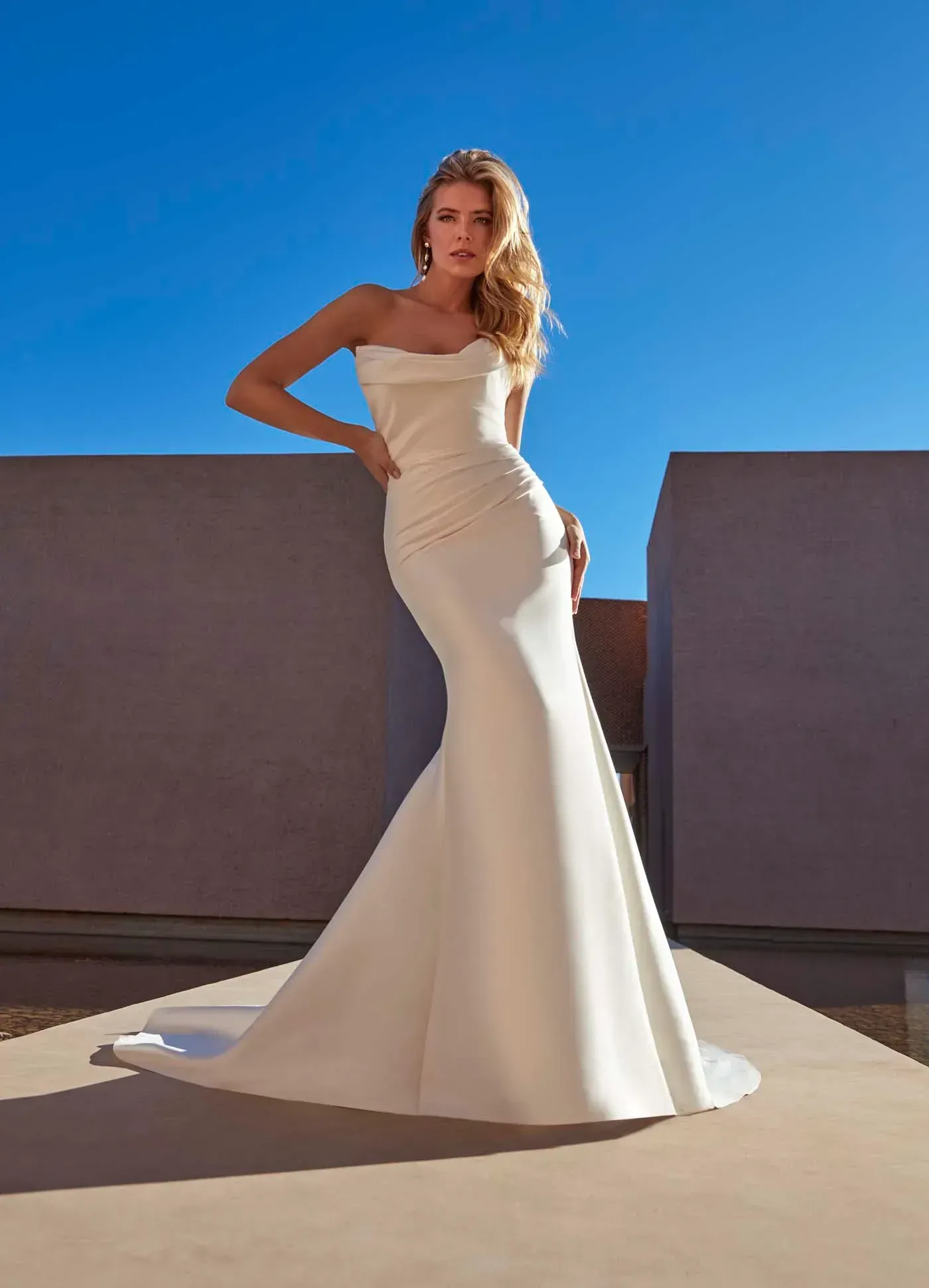 Woman in a white mermaid wedding dress, standing on a terrace with blue sky background.