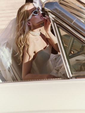 Woman in a white wedding dress holds a sheer veil, looking at the camera. She has long hair and gold earrings.