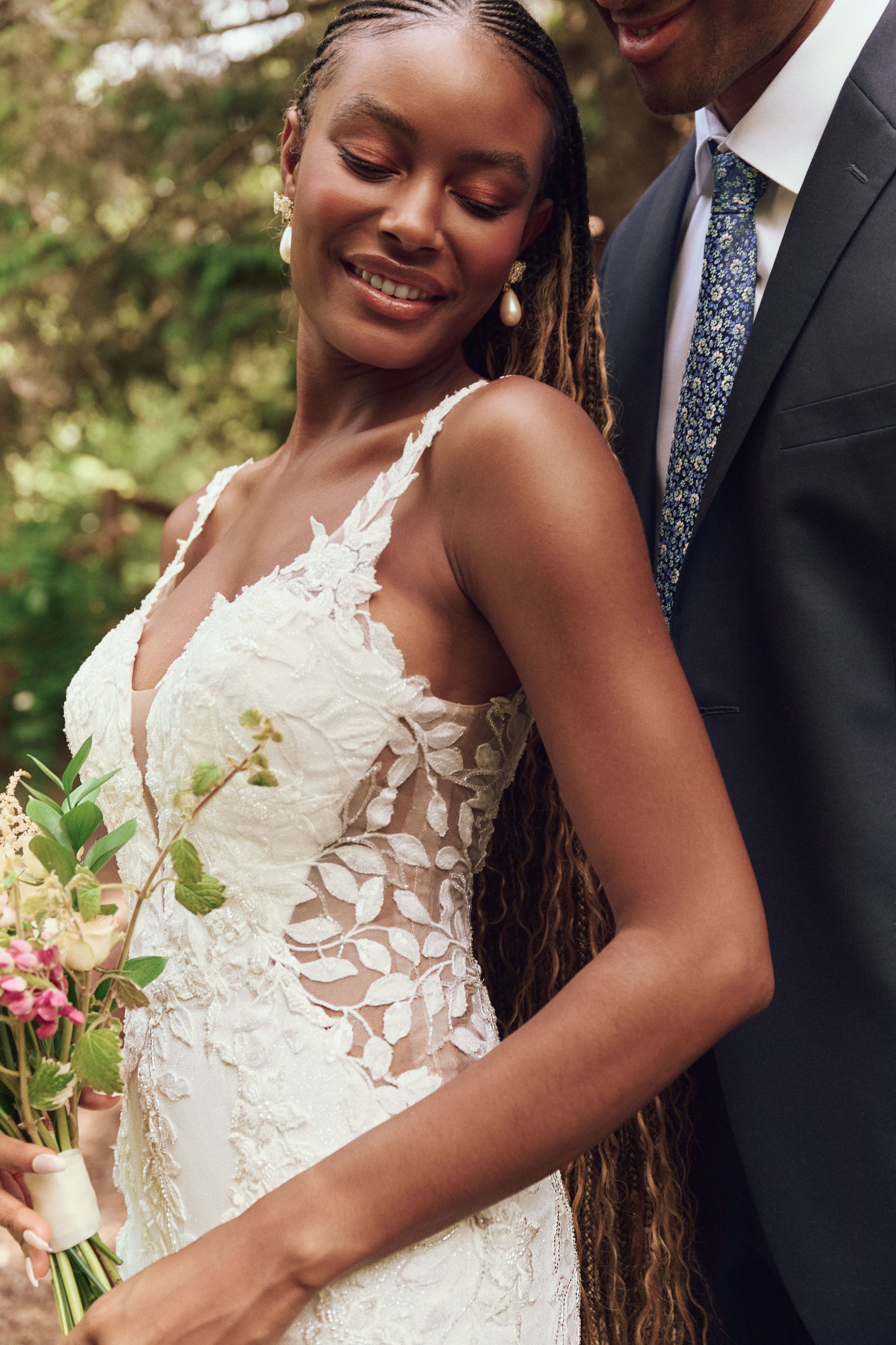 Smiling bride in white strapless gown and gold jewelry, holding flowers outdoors in front of a column.