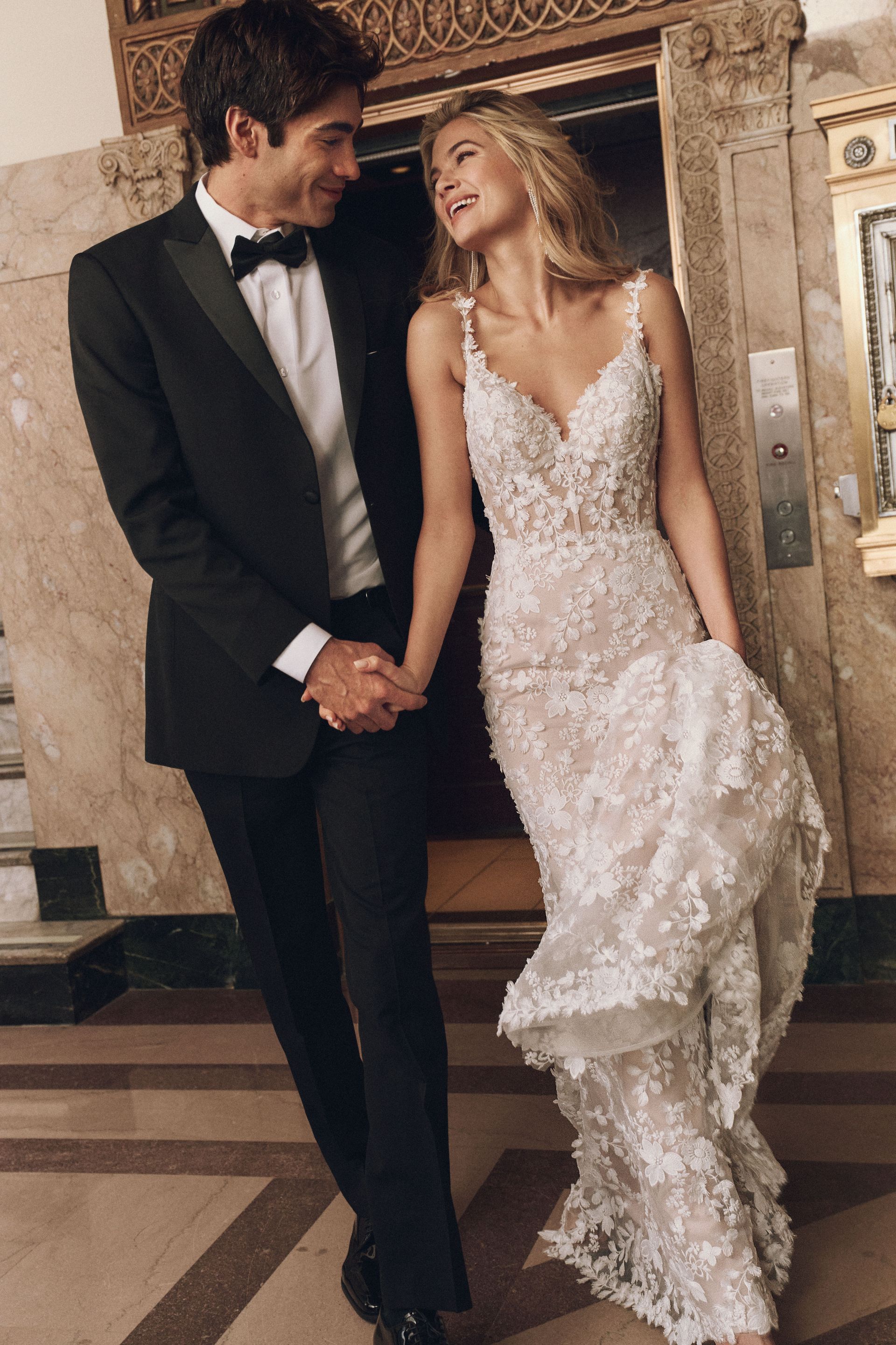 Groom in a tuxedo and bride in a floral gown holding hands, smiling, in an ornate building.