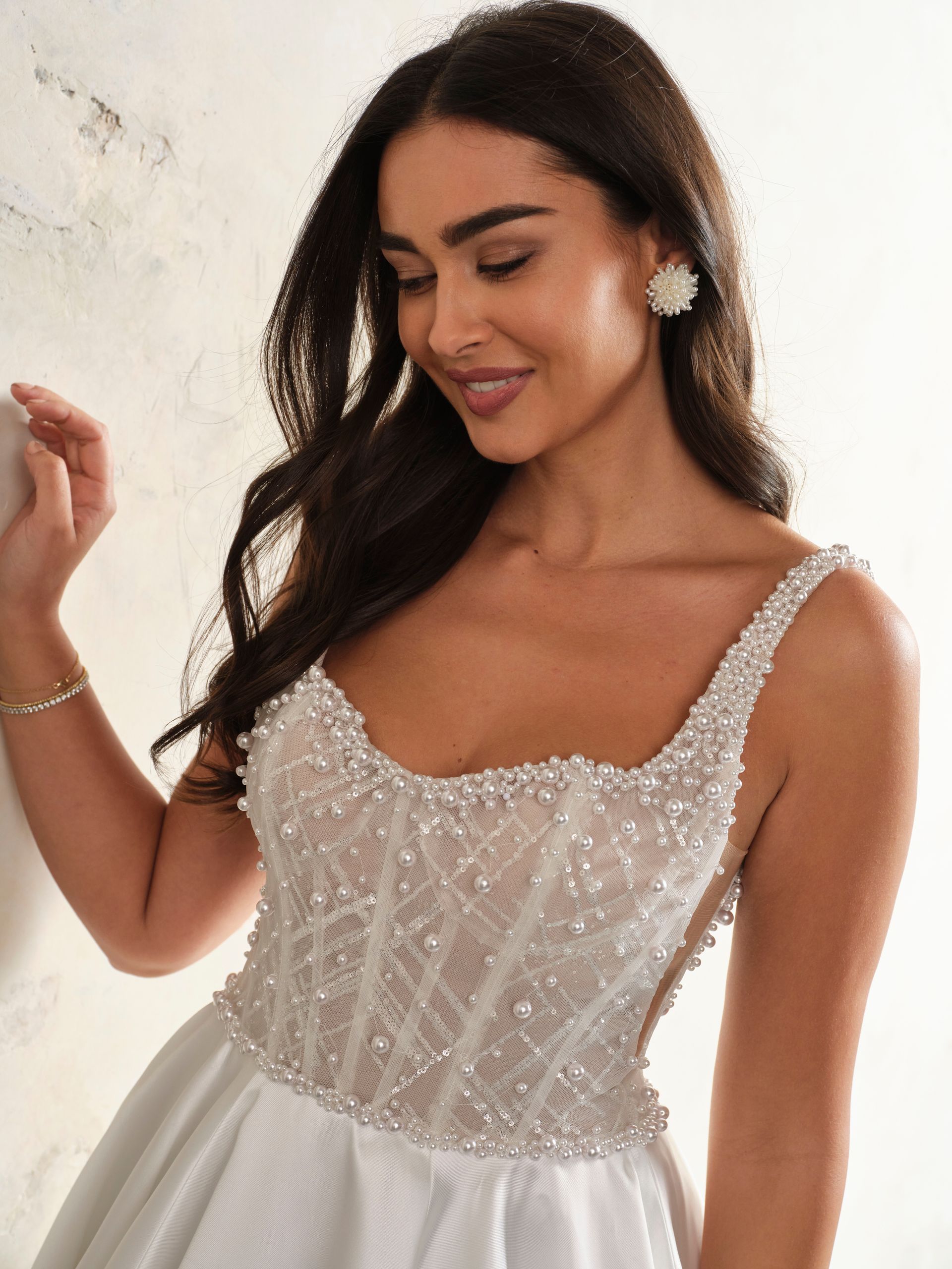 Woman in a white beaded gown, smiling, indoors, next to a light-colored wall.