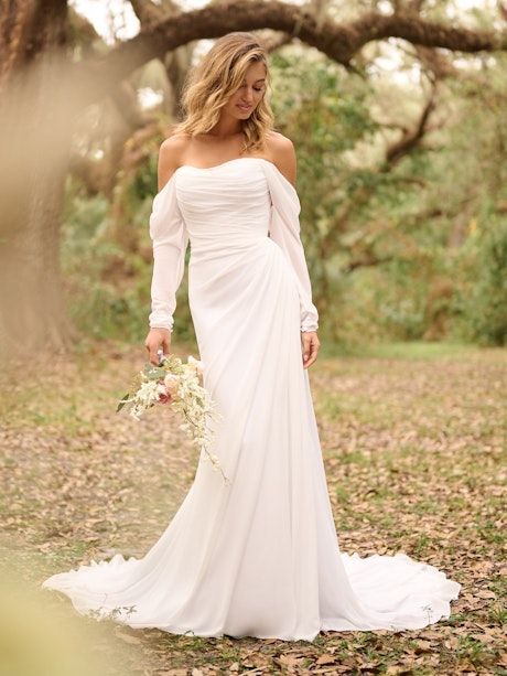 Blonde woman in white off-the-shoulder wedding dress holding flowers, outdoors in a wooded area.
