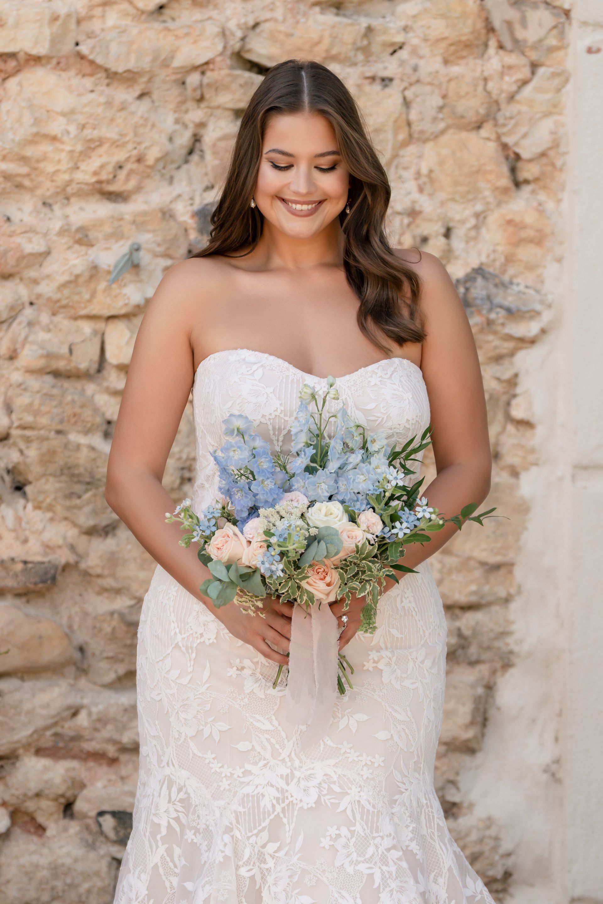 Woman in white strapless gown poses in a room with dark wood paneling and a tiled floor. She wears gold earrings and touches her hair.