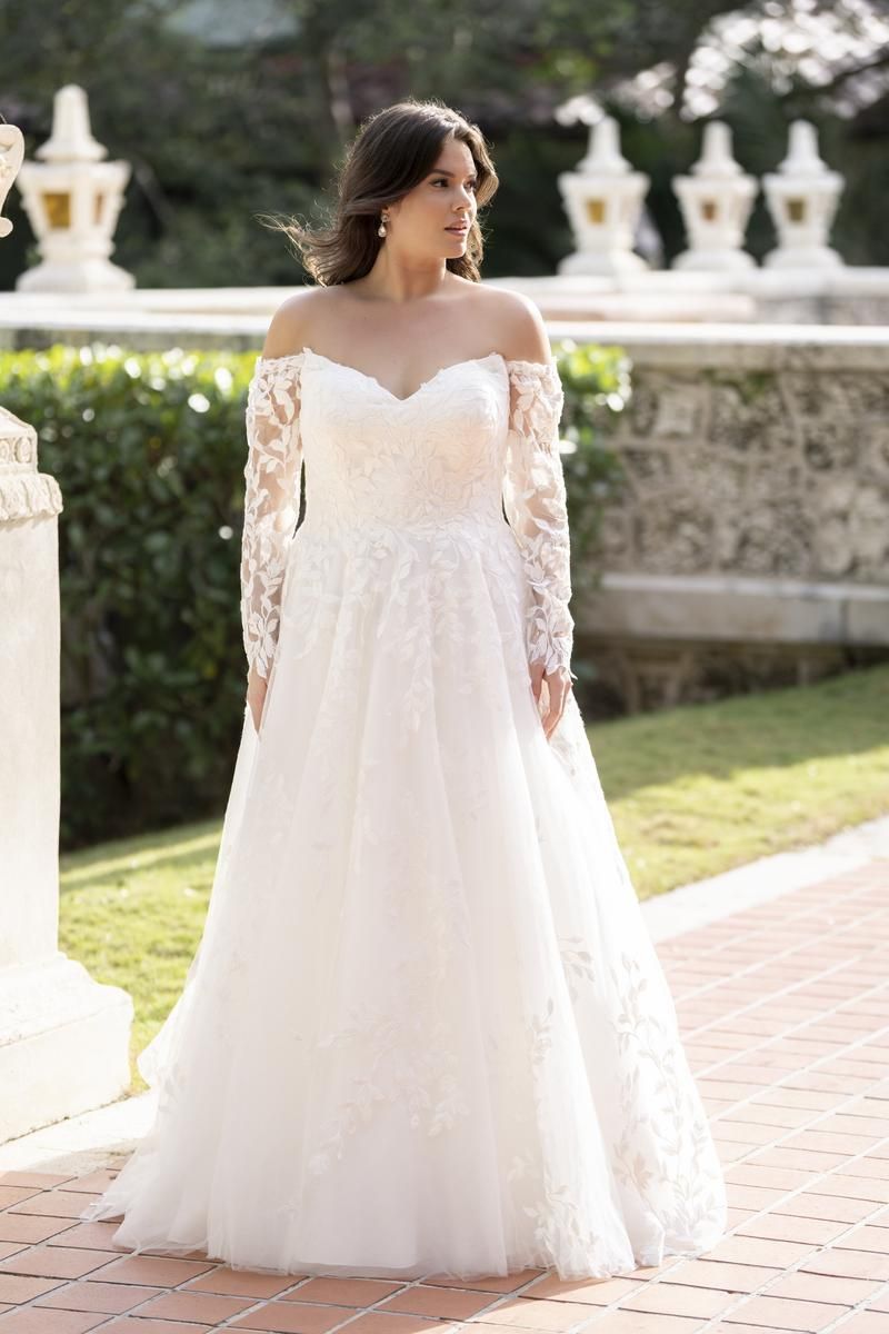Woman in a white off-the-shoulder wedding dress stands outdoors on a brick path.
