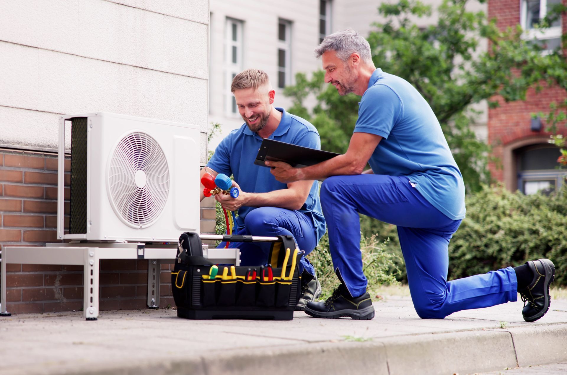 Two technicians kneel beside an outdoor HVAC unit, checking tools on a concrete patio.