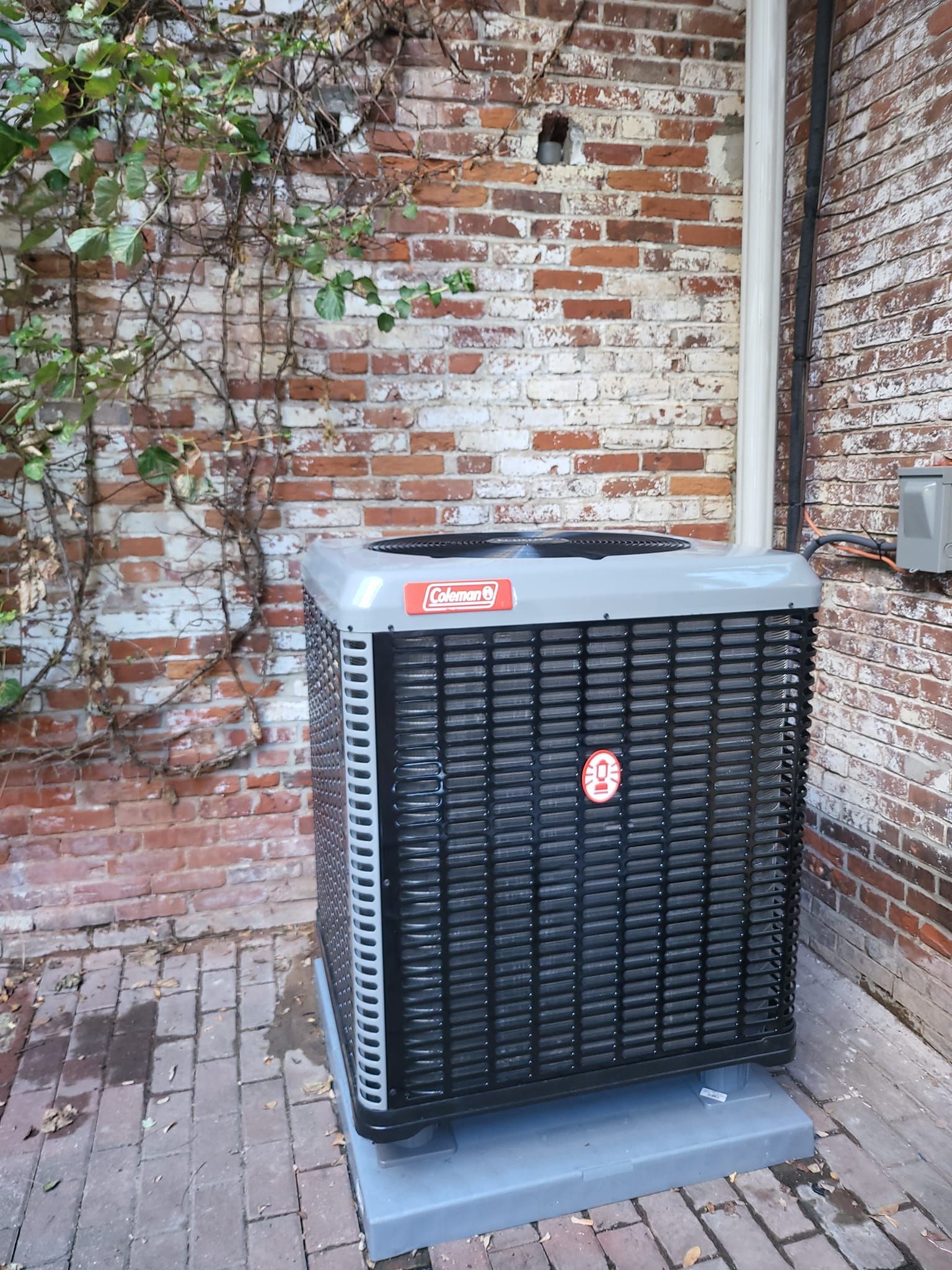 A black air conditioner is sitting in front of a brick wall.