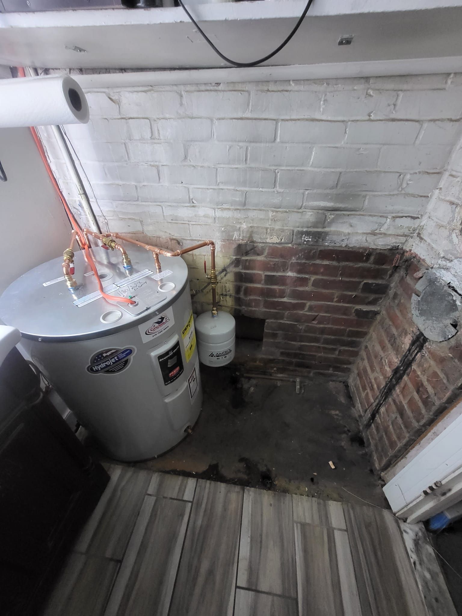A water heater is sitting in a corner of a room next to a brick wall.