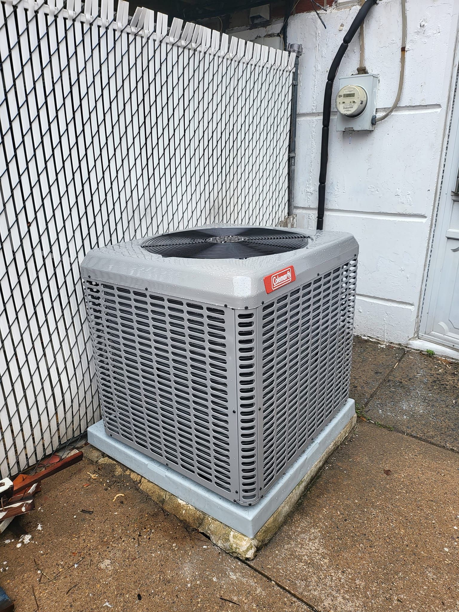 An air conditioner is sitting on the sidewalk next to a fence.