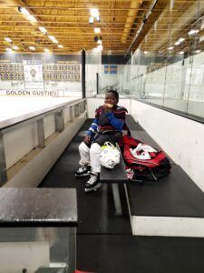 Boy in ice skates, shoulder pads, eating while sitting on a bench at an ice rink. Hockey gear nearby.
