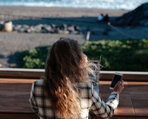 Woman with long hair in plaid jacket uses phone at a table, beach in background.