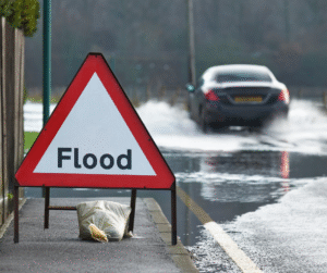 Flood warning sign on roadside; car driving through flooded street.