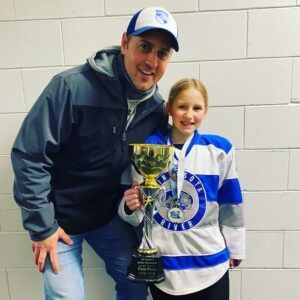 Man and girl in hockey jerseys holding a trophy. Both smiling in front of a white wall.