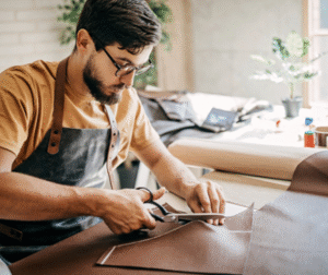 Man wearing glasses and an apron cuts leather with scissors at a workbench.