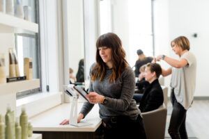 Woman in a salon looking at a tablet, another stylist cutting someone's hair in the background. White and bright space.