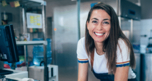 Woman smiles broadly in a kitchen setting. She wears a white shirt, apron, and appears to be working.