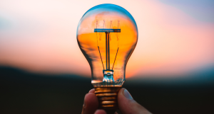 Hand holding a lit lightbulb against a sunset sky.