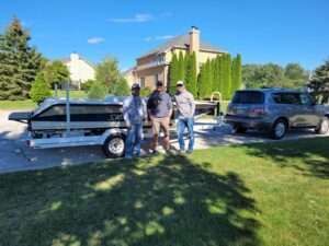 Three men stand next to a boat on a trailer, parked on a paved area with a house and SUV in the background.
