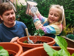 Man and girl watering plants in terracotta pots; girl smiles as she pours water.