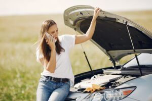 Woman on phone by car with open hood, stranded in field.