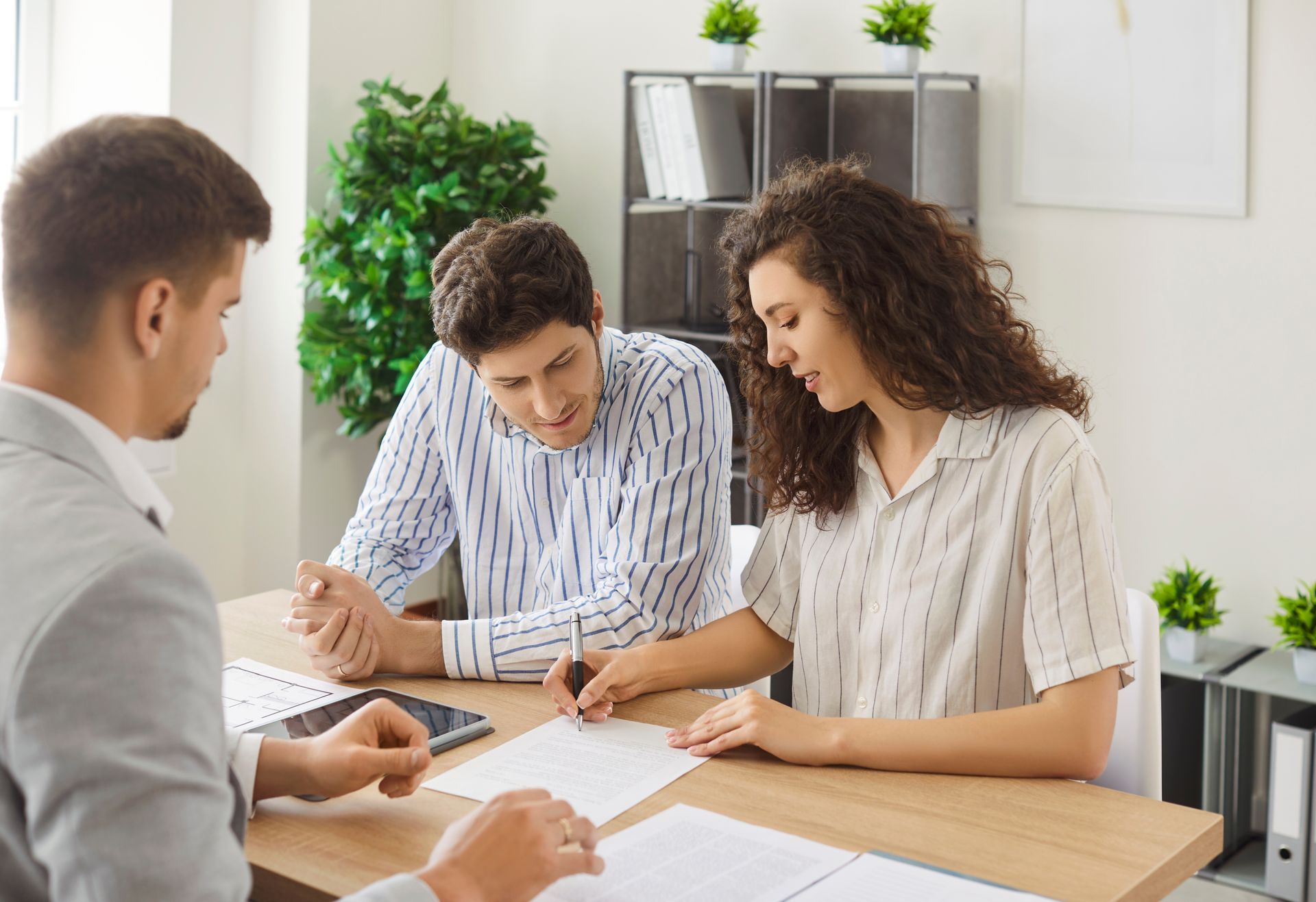 Two people sign documents at a table with an advisor. Office setting, neutral colors.