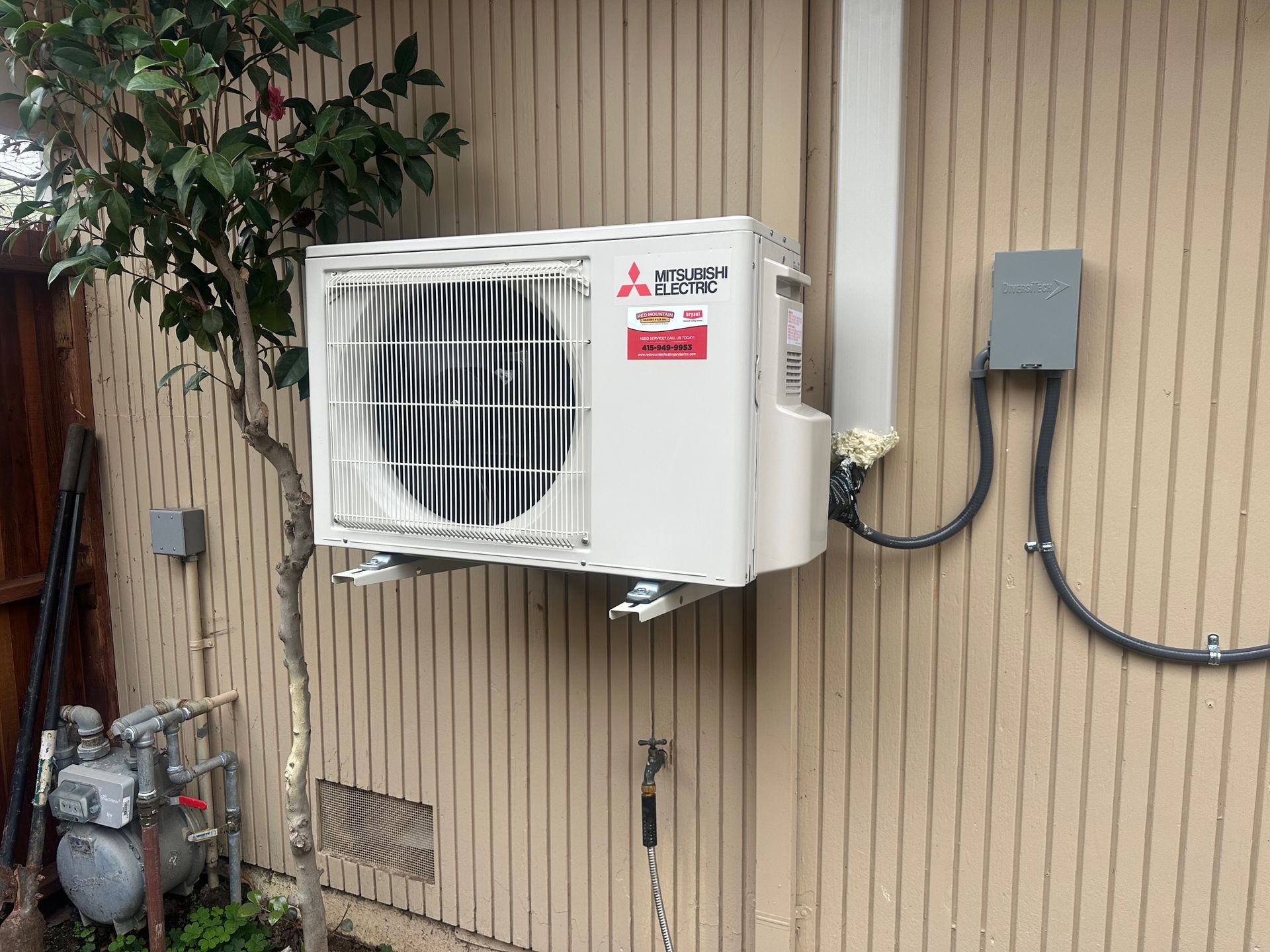 White Mitsubishi air conditioning unit mounted on a beige building exterior with electrical conduit and a small tree.