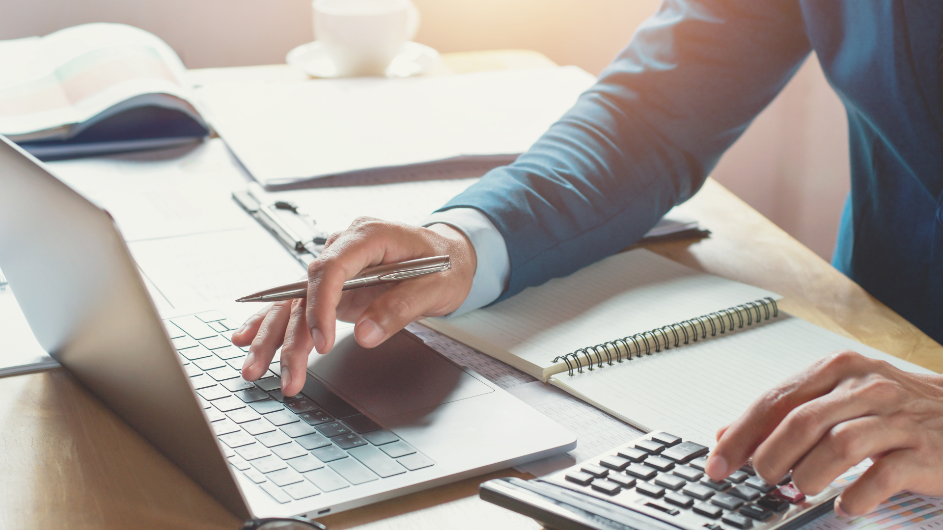 Hands typing on a laptop beside an open notebook and coffee on a bright desk