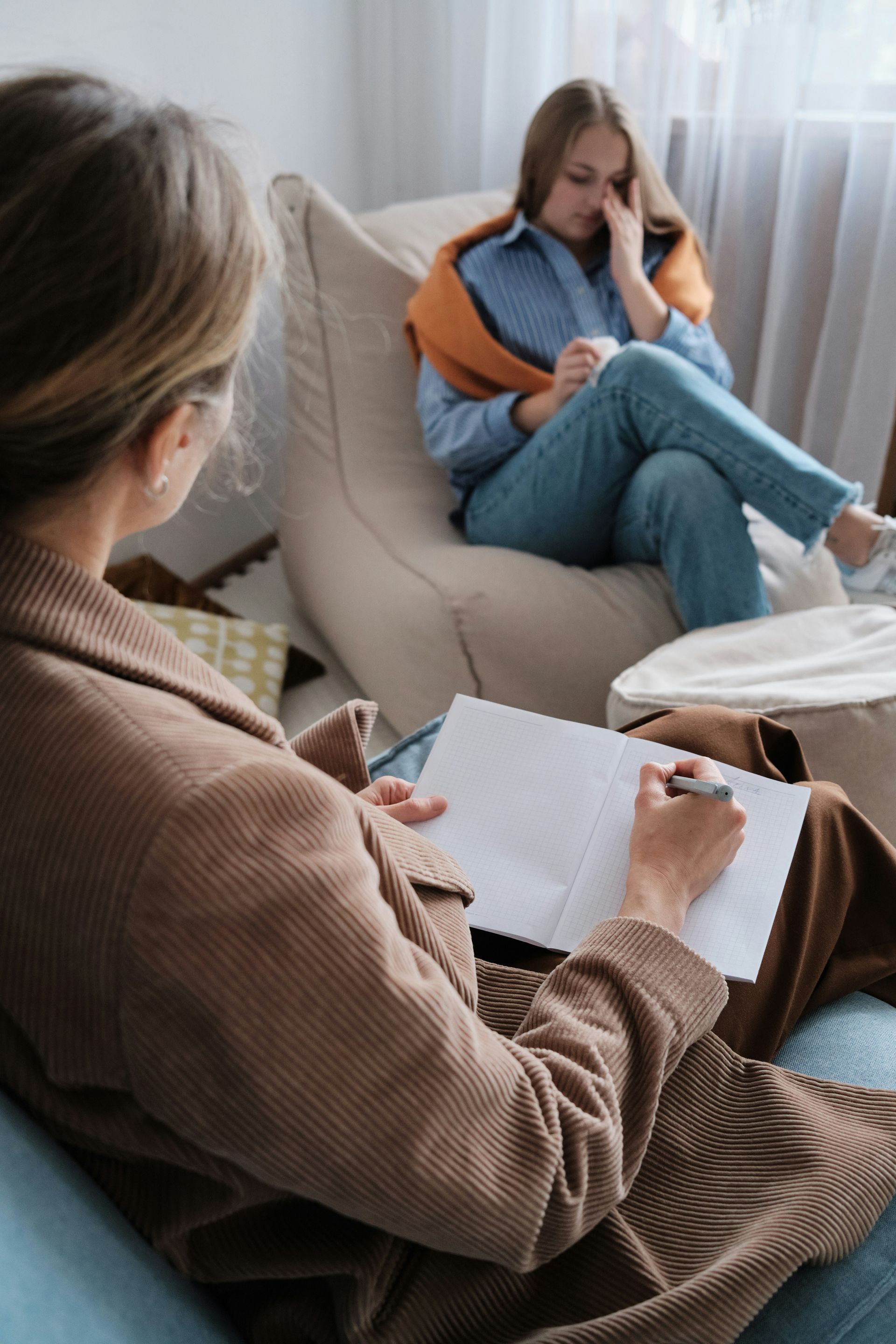 A woman is sitting in a chair talking to another woman while a woman writes in a notebook.