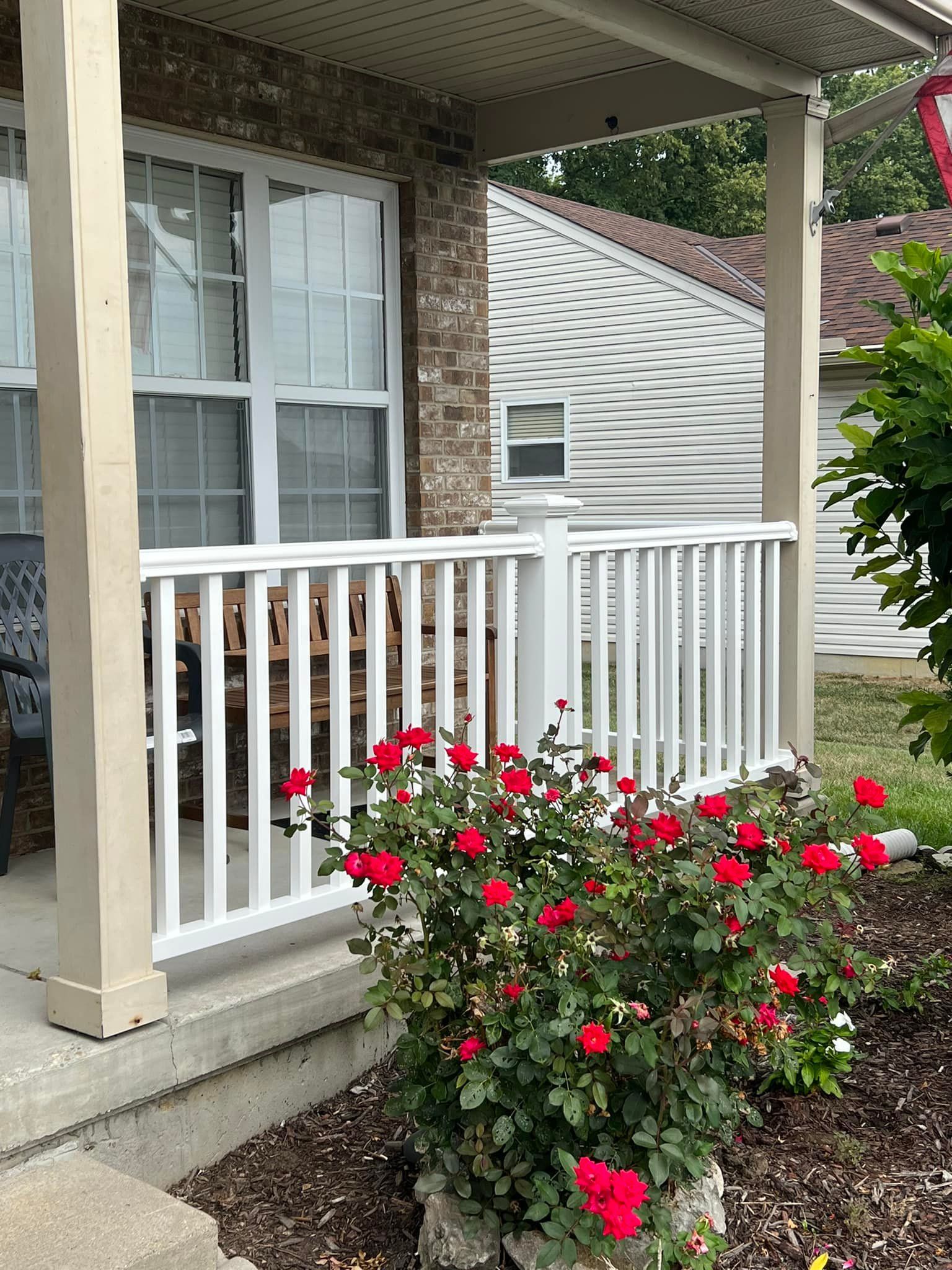White-railed porch with red roses; adjacent window, brown brick. Tan posts frame the view of the porch.