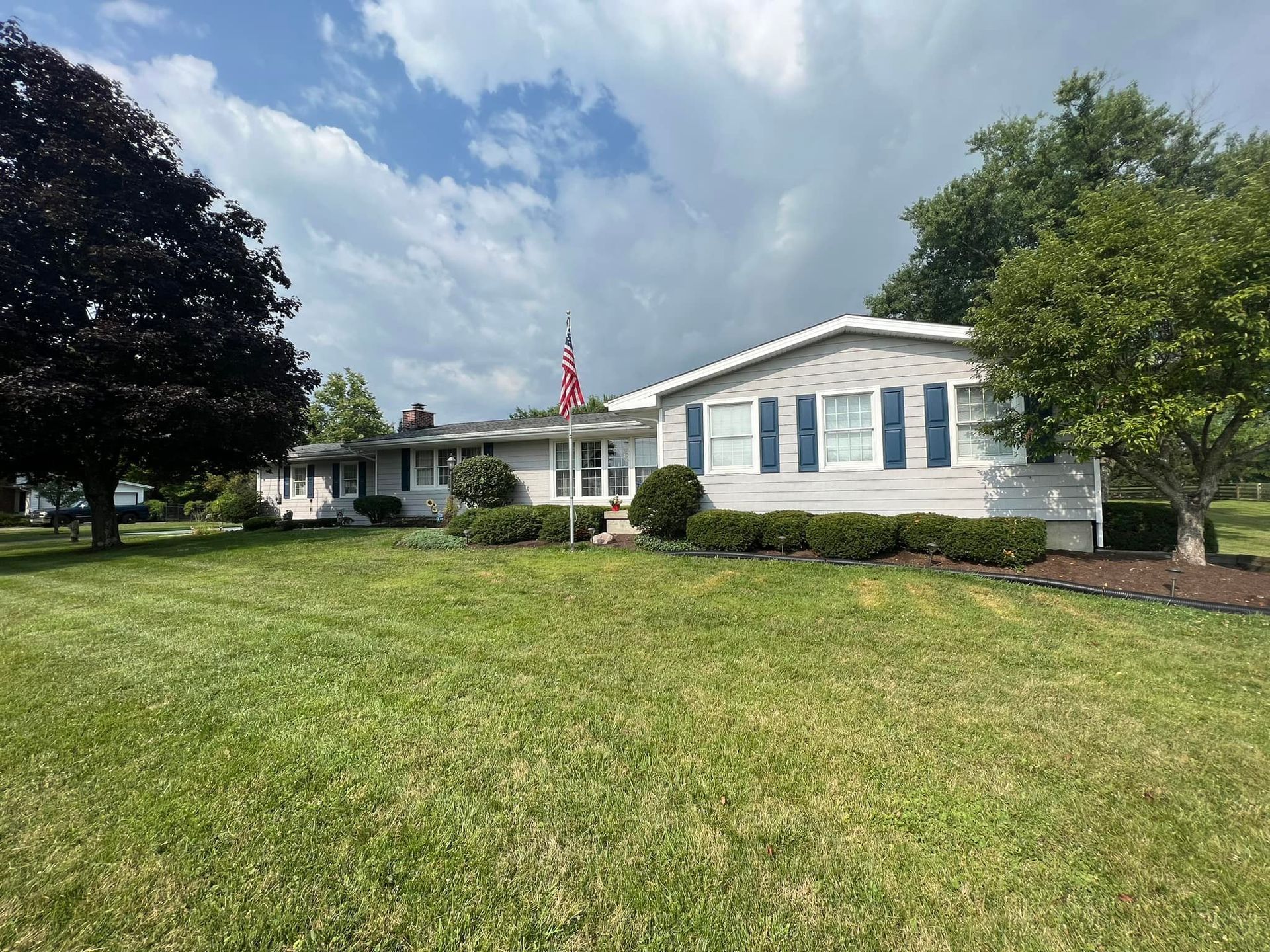 Ranch-style house with blue shutters, American flag, and green lawn under a cloudy sky.