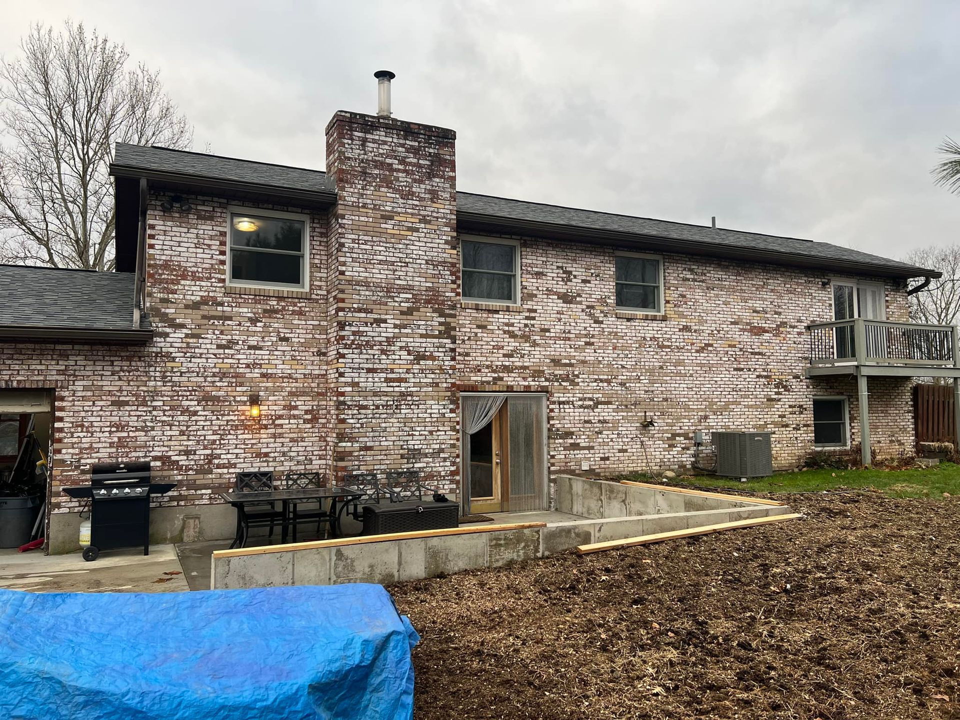 Brick house exterior with chimney, windows, and small deck. Concrete foundation visible, covered by leaves.