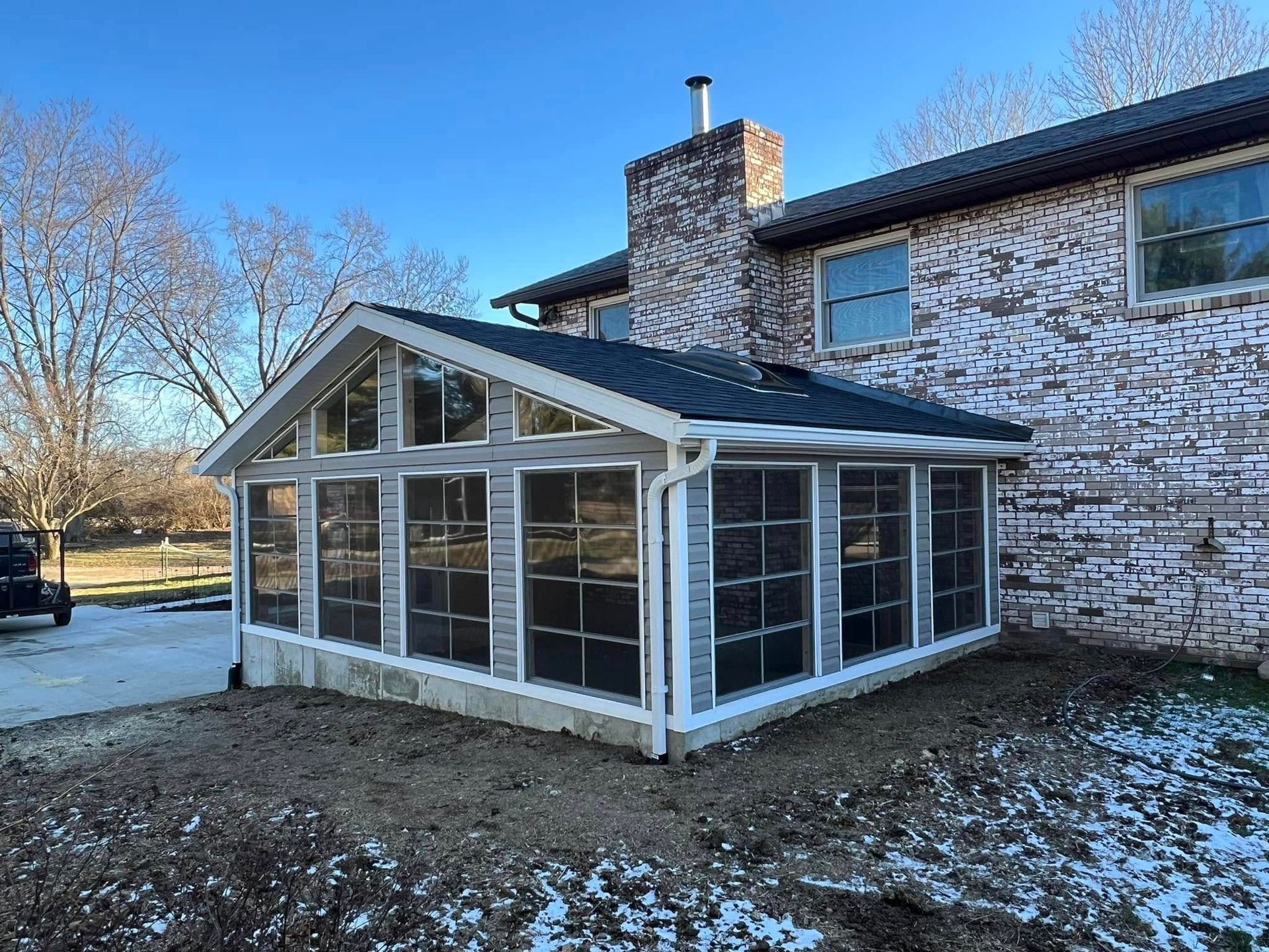 Sunroom addition with gray siding and roof attached to a brick house.