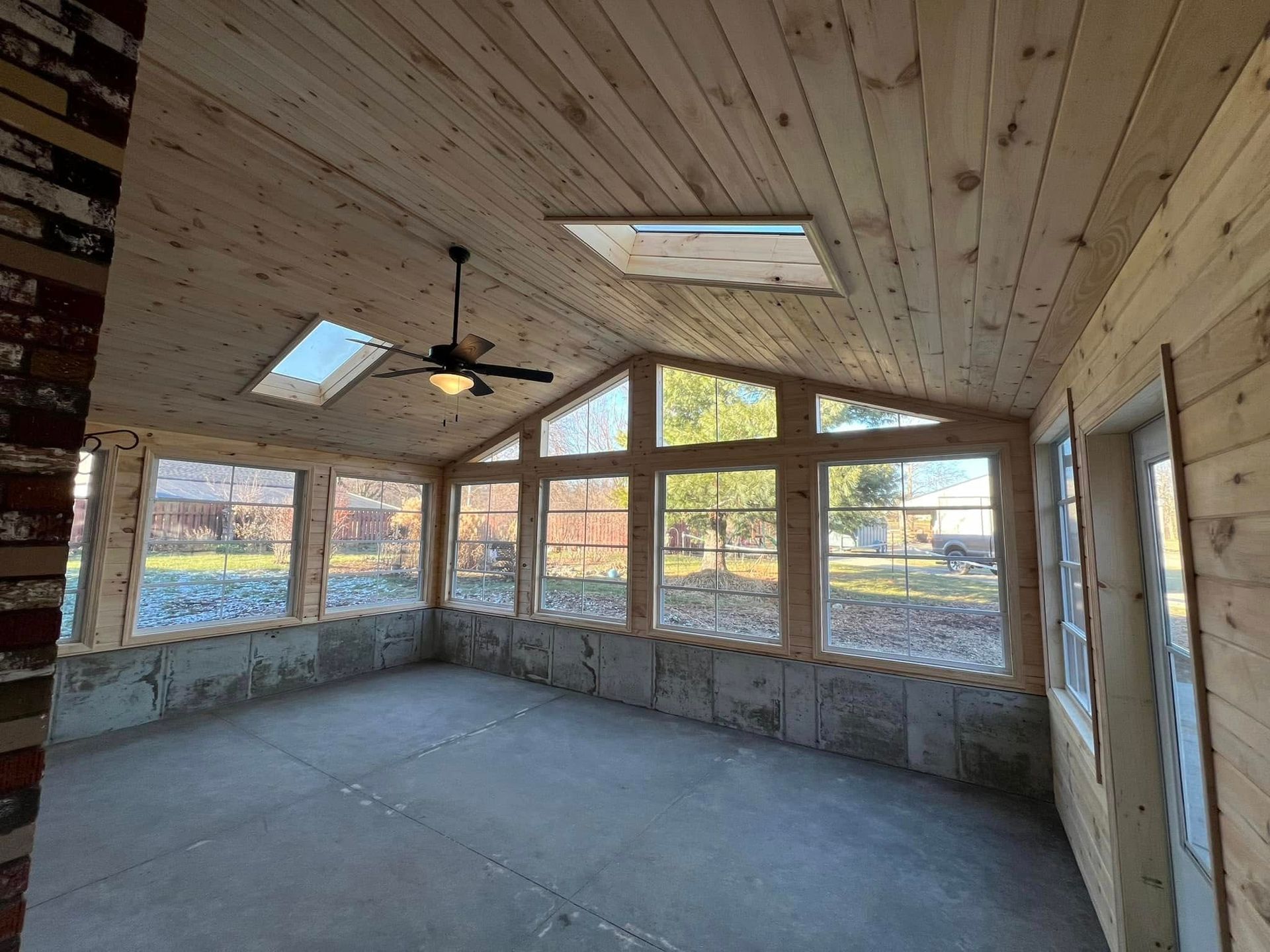 Sunroom with wooden ceiling and walls, large windows, two skylights, ceiling fan, and concrete floor.