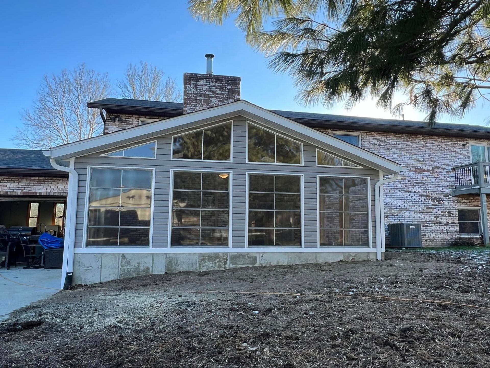 Sunroom addition with large windows and gray siding, attached to a brick house on a sunny day.
