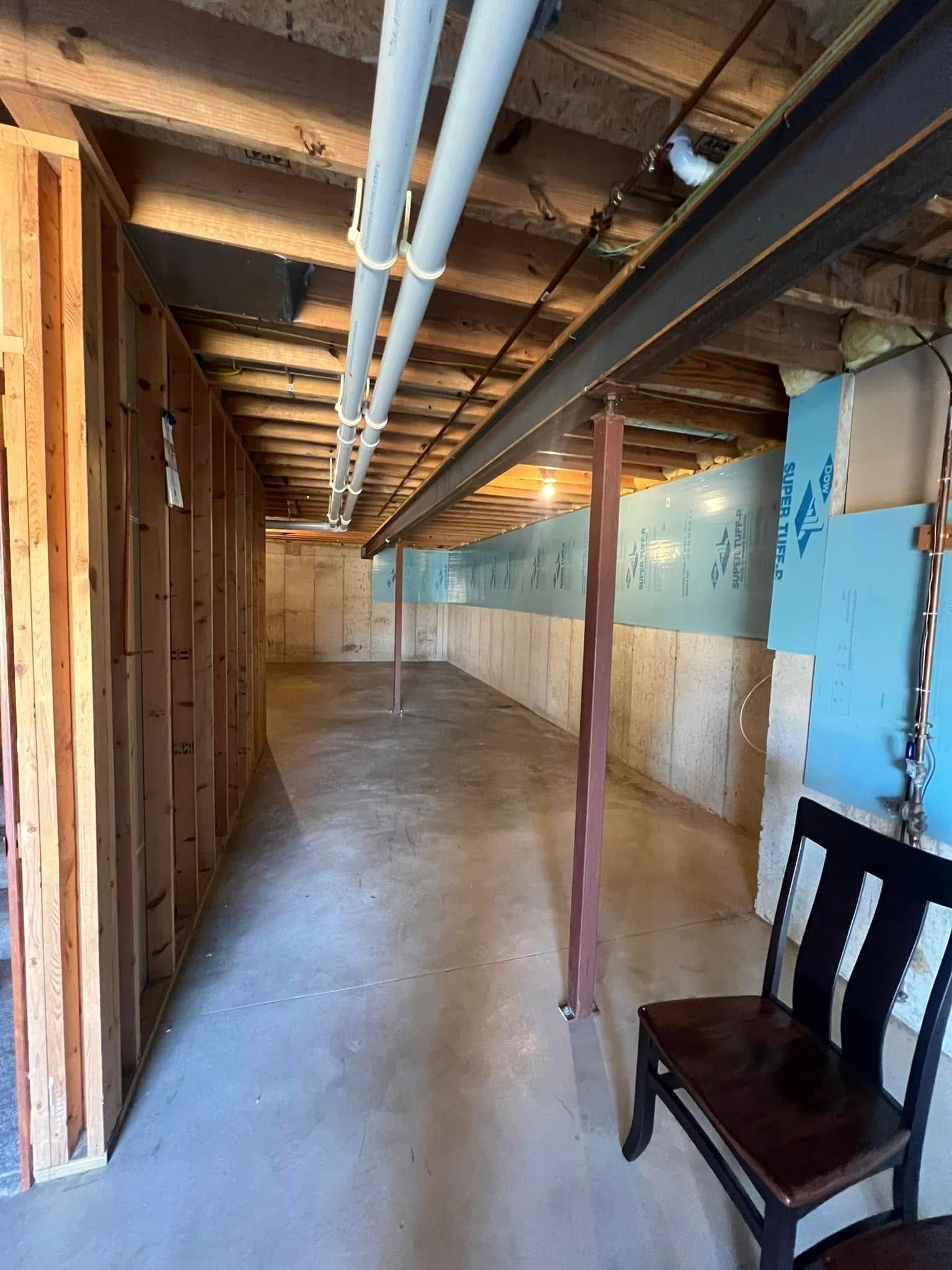 Unfinished basement with concrete floor, exposed framing, and pipes running along the ceiling. A chair sits on the right.