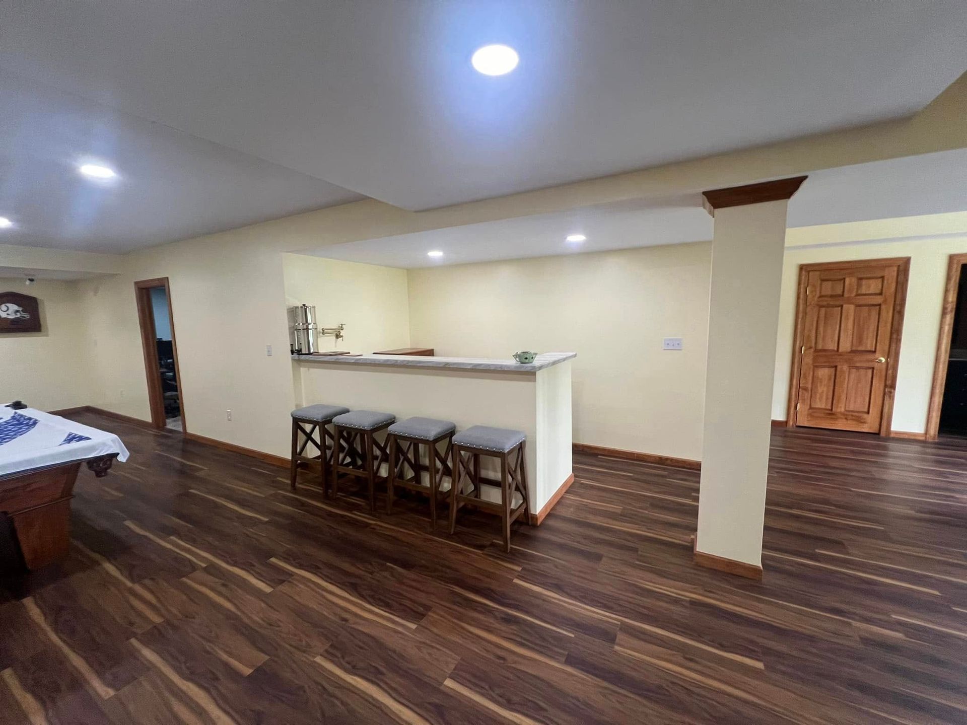 Basement with bar, stools, wooden floor, and a door, lit by recessed lighting.