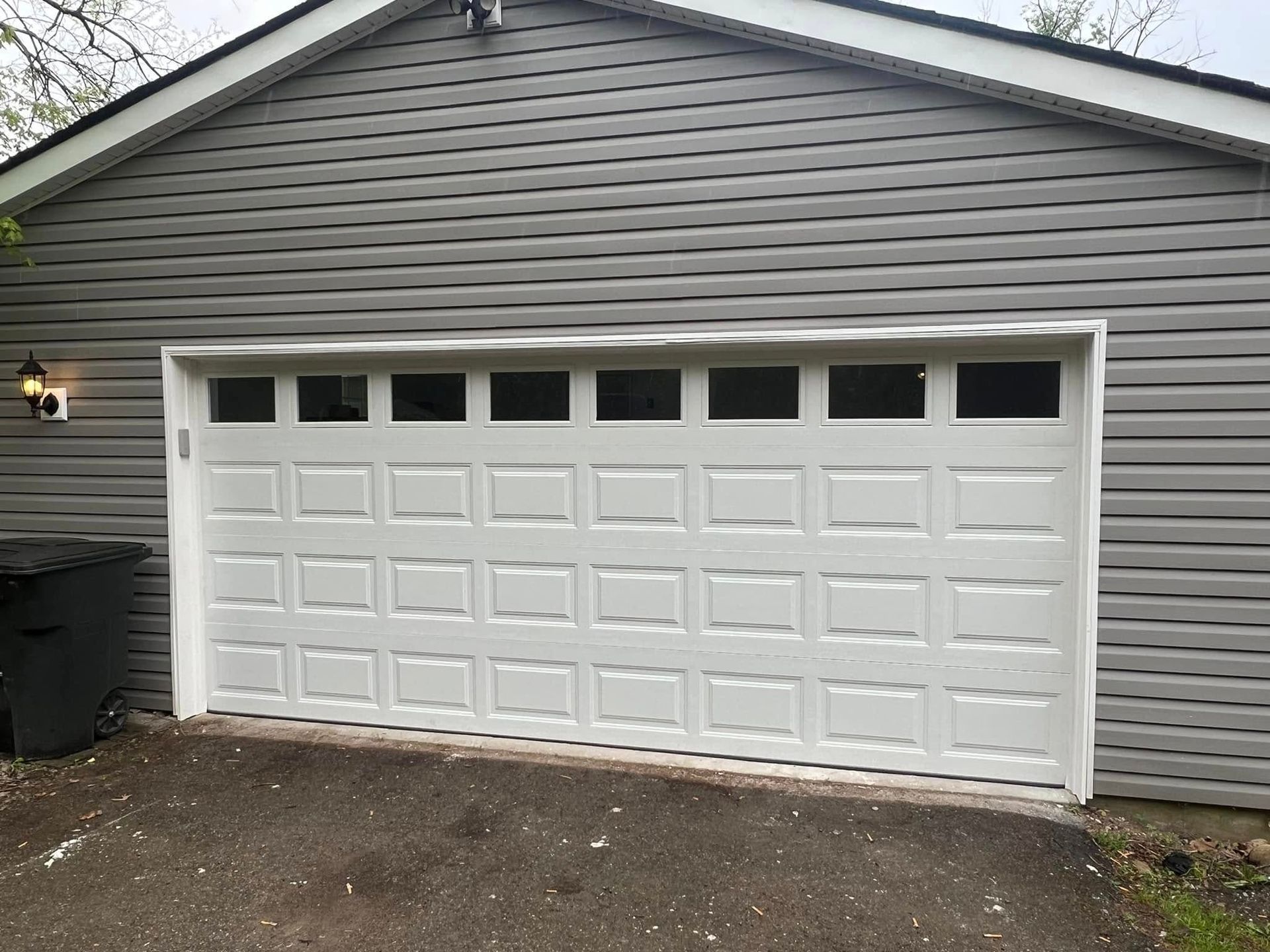 White garage door with windows, in front of a gray siding garage, next to a trash can.