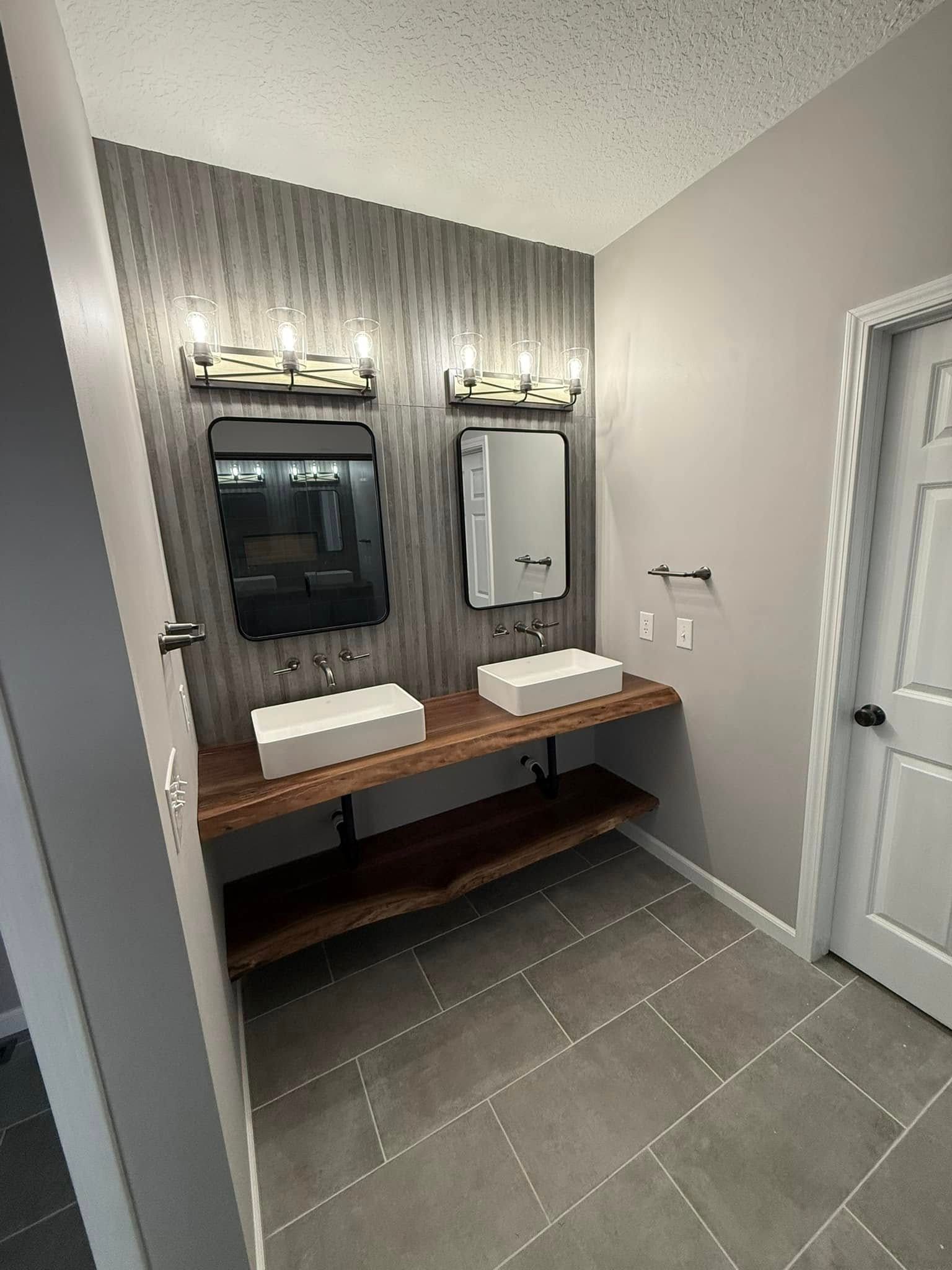 Bathroom with dual sinks on a wooden shelf, gray tiled floor, and textured wall behind mirrors.