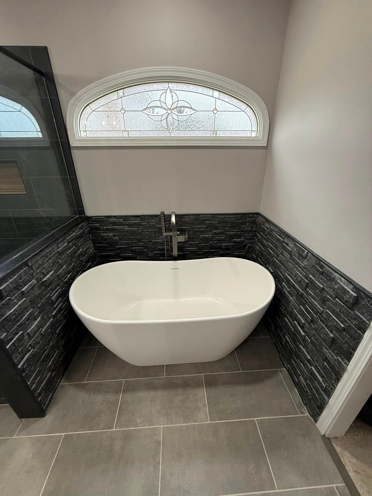 Freestanding white bathtub under arched window in a bathroom with dark tiled accent walls and gray tiled floor.